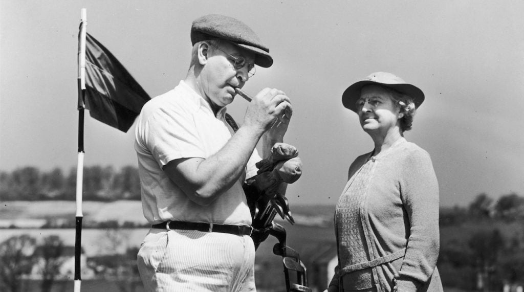 irca 1945: Full-length image of an elderly man and woman standing on a golf course beside the pin, as the man lights a cigar. The woman stands with a bag of clubs. (Photo by Harold M. Lambert/Lambert/Getty Images)