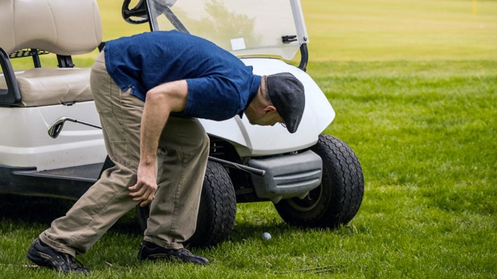 A man looks for his golf ball under a cart.