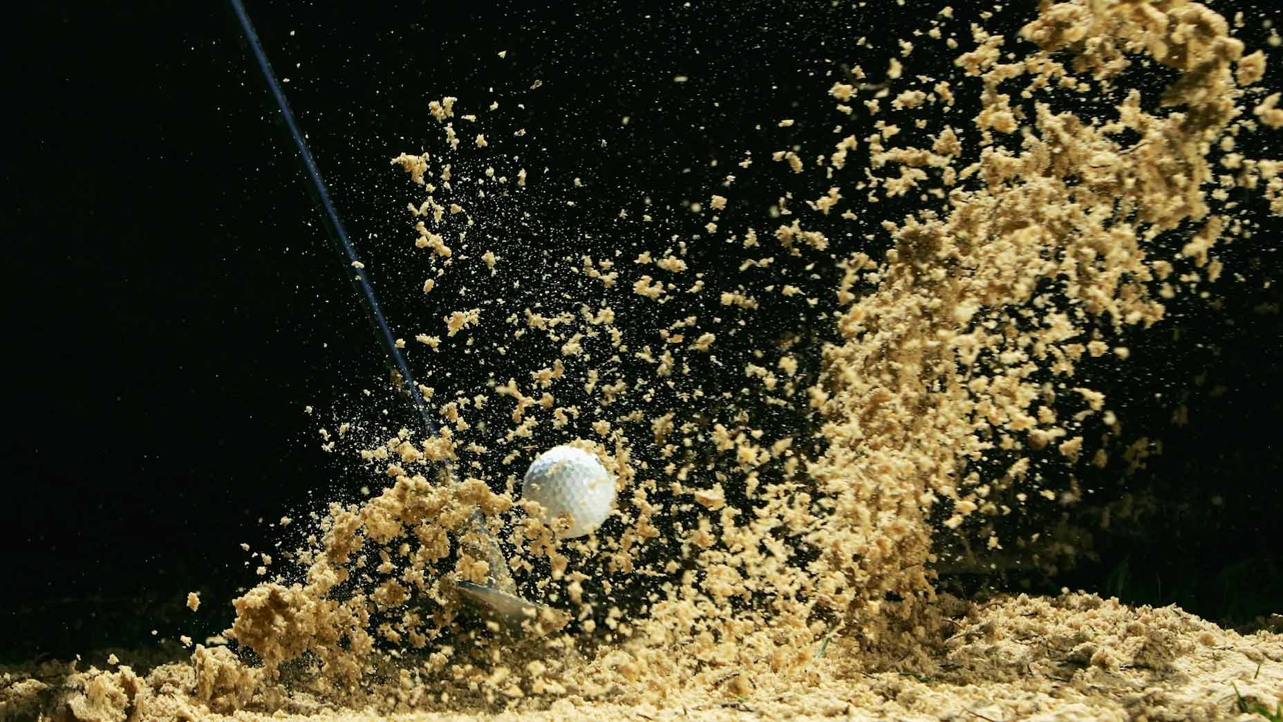 Sand flies up as a golf ball in a bunker is struck by a wedge in front of a black background