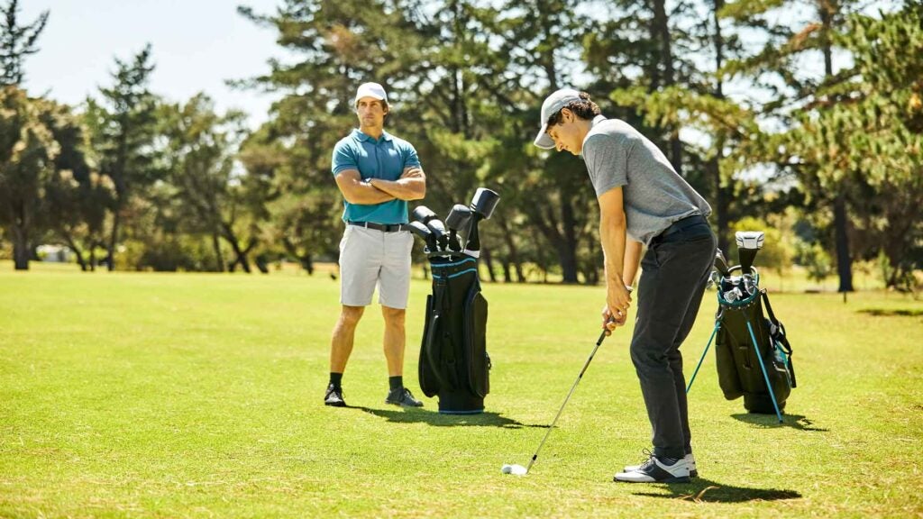 stock image of golfer practices chip shot next to other golfer standing with arms crossed