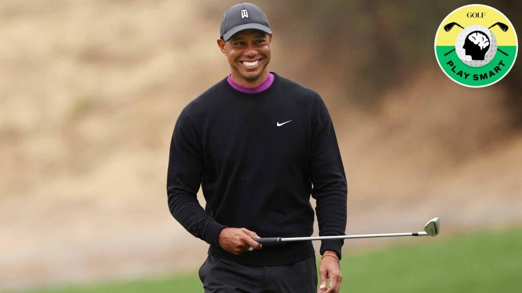 tiger woods holds a club in his right hand and smiles while wearing a black nike sweater