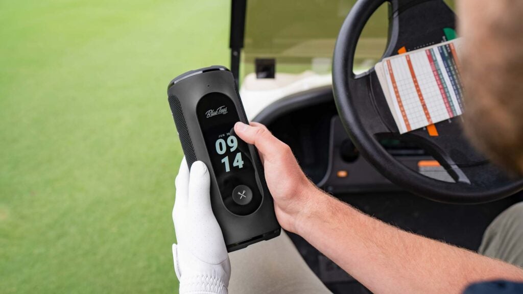 A golfer holds a Blue Tees Player+ GPS speaker while sitting in a golf cart