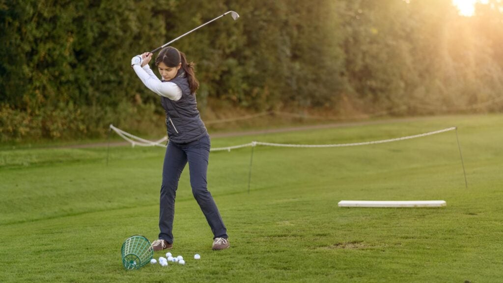 Female golfer standing on the driving range on a late afternoon day, concentrating while swining her club