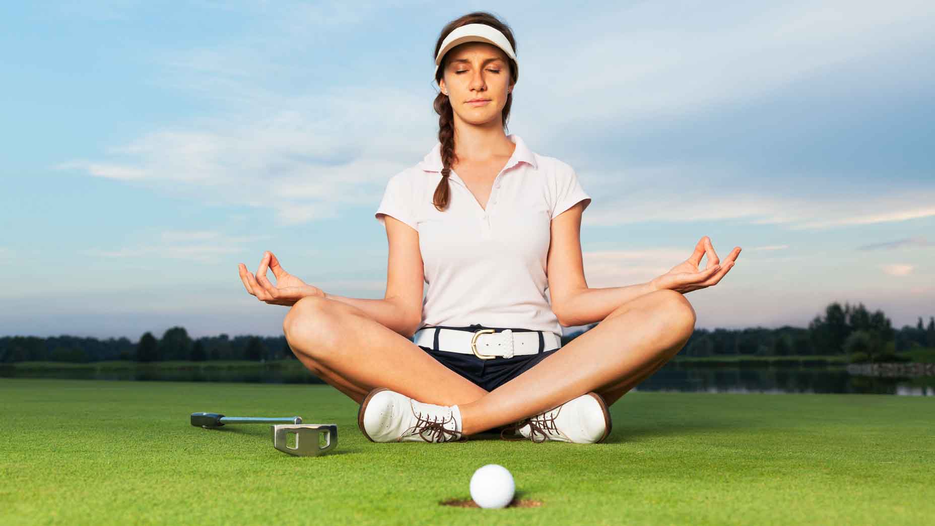 Woman golf player sitting on green in yoga posture focusing on ball dropping into cup.