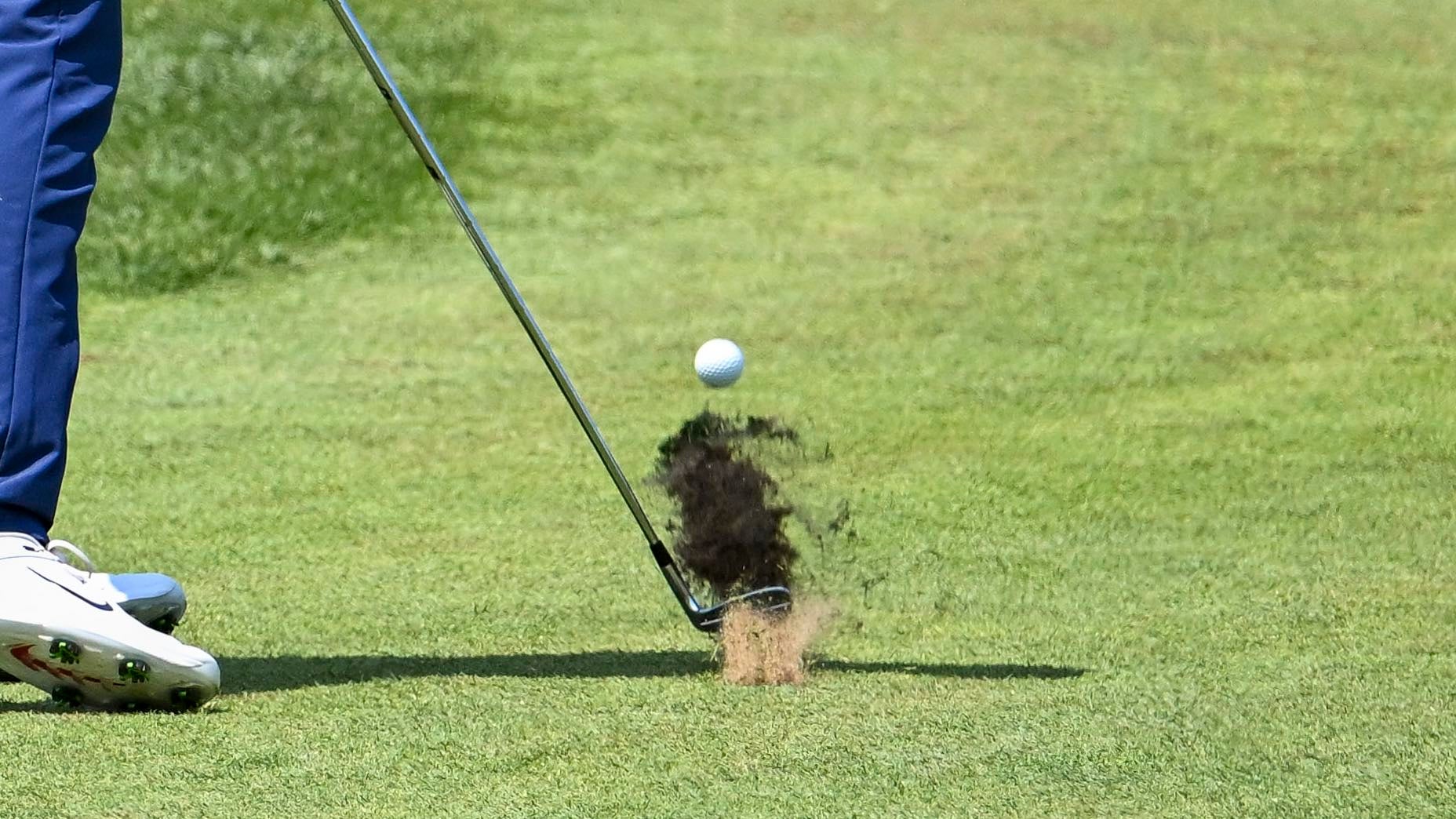 Golfer hits ball on golf course causing divot to fly in air.