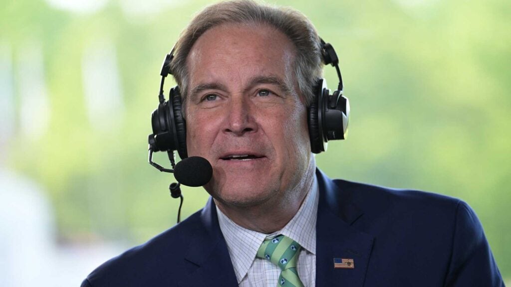 jim nantz speaks into headset at the memorial tournament in a blue suit with green tie