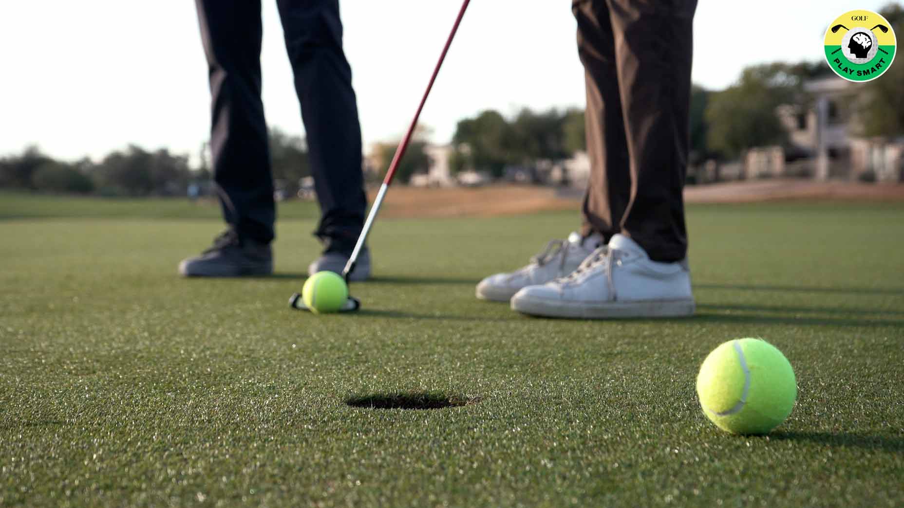 person stands on green rolling tennis ball with putter while another person stands behind