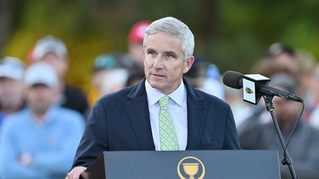 jay monahan stands at a podium at the presidents cup in suit and green tie