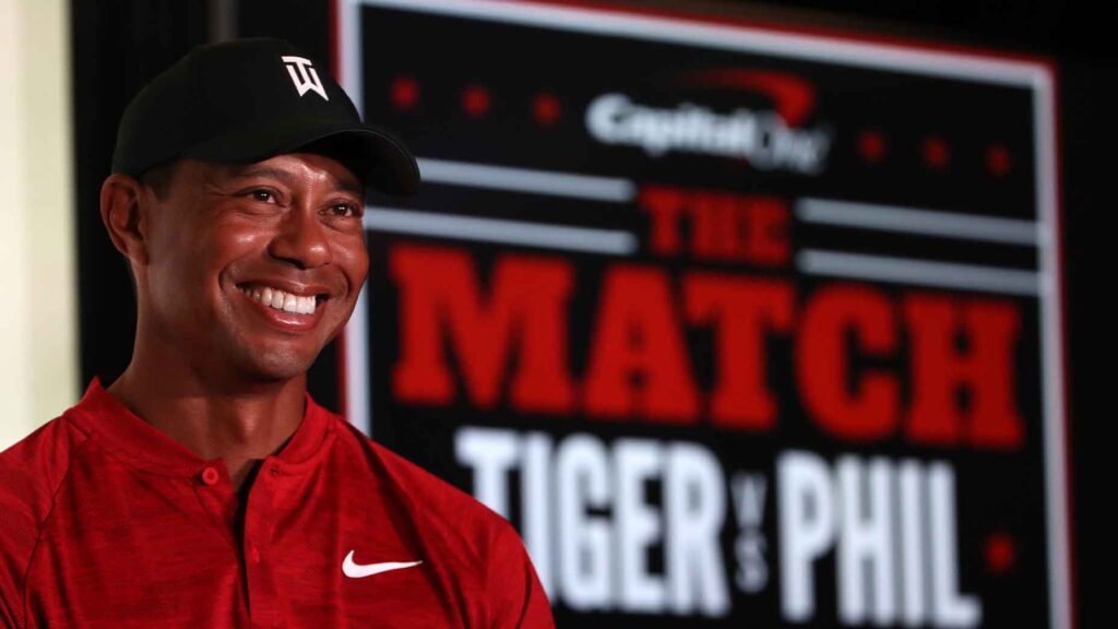 tiger woods smiles in front of the match tiger vs phil signage in red mock neck shirt