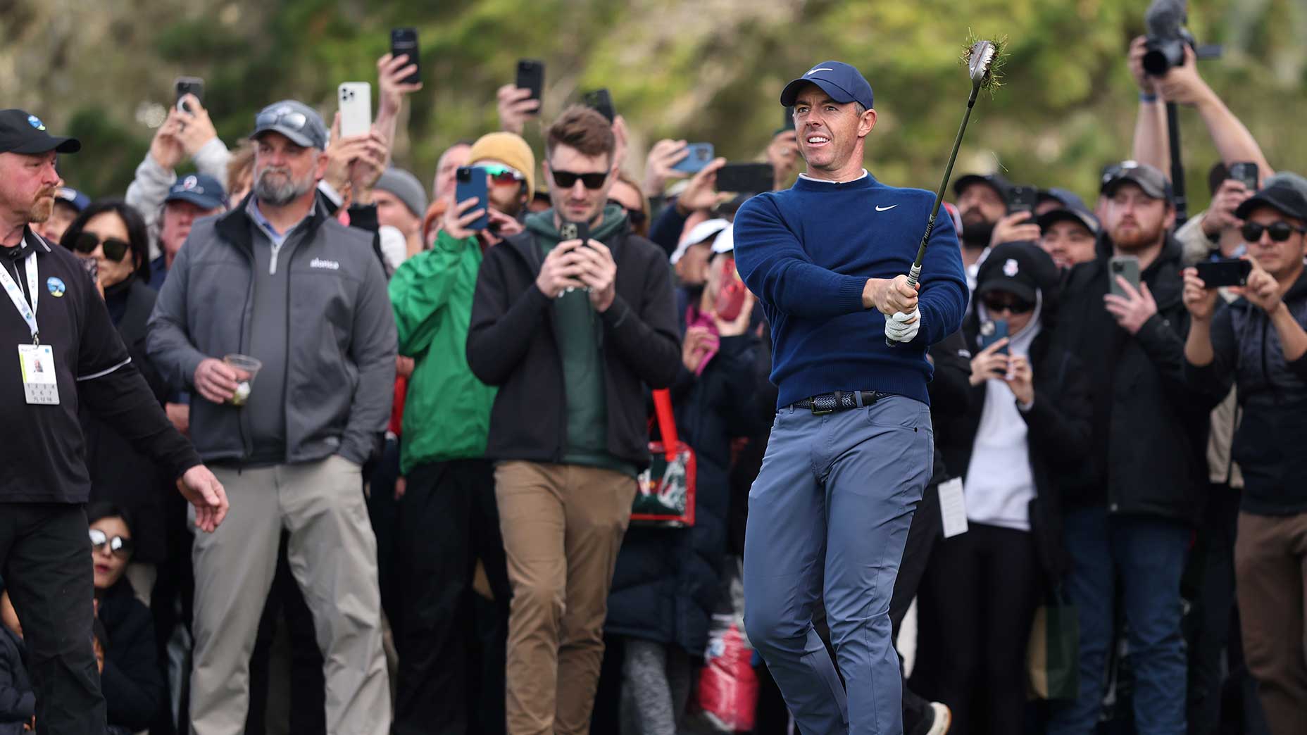 rory mcilroy swings iron in front of gaggle of fans in navy sweater and slate pants