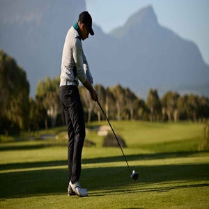 golfer in white shirt and black pants hits golf shot with mountains in background