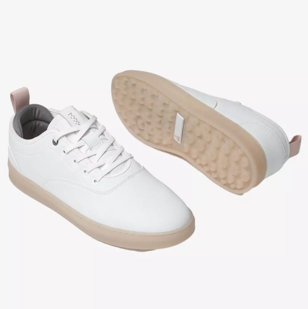 A pair of white low-top sneakers with beige soles, white laces, and a light pink pull tab at the heel&mdash;perfect as contenders for the best women's golf shoes 2026. One shoe is upright, and the other is turned to show its textured sole.