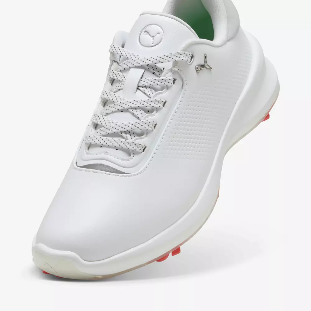 A white athletic shoe with a textured side panel, patterned laces, and a silver Puma logo on the tongue and upper side&mdash;one of the best golf shoes 2026&mdash;features a low-cut design and a light pink sole with visible spikes.
