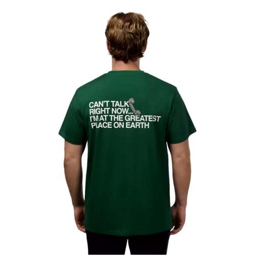 A person with light brown hair, seen from behind, wears a dark green T-shirt featuring one of the best looks from The First Major. The back reads: CANT TALK RIGHT NOW. IM AT THE GREATEST PLACE ON EARTH, with a small golf tee graphic.