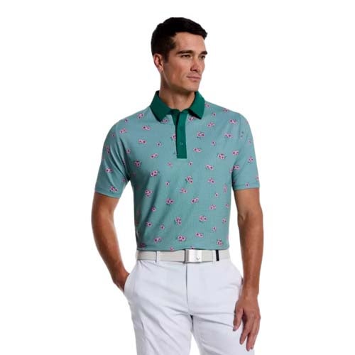 Man wearing a light teal short-sleeved polo with a green collar and pink flamingos, paired with white pants, stands with one hand in his pocket—channeling some of the best looks from The First Major.
