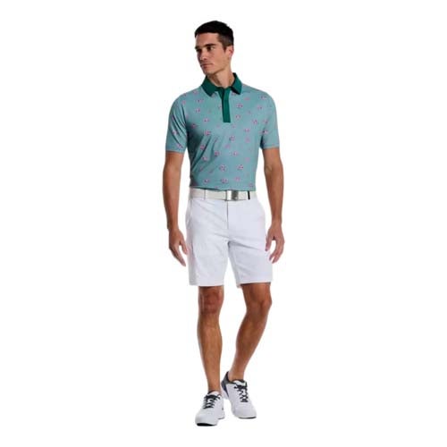 A man stands wearing a teal patterned polo, white shorts, a white belt, and white golf shoes with black accents—channeling one of the best looks from The First Major—against a plain white background.