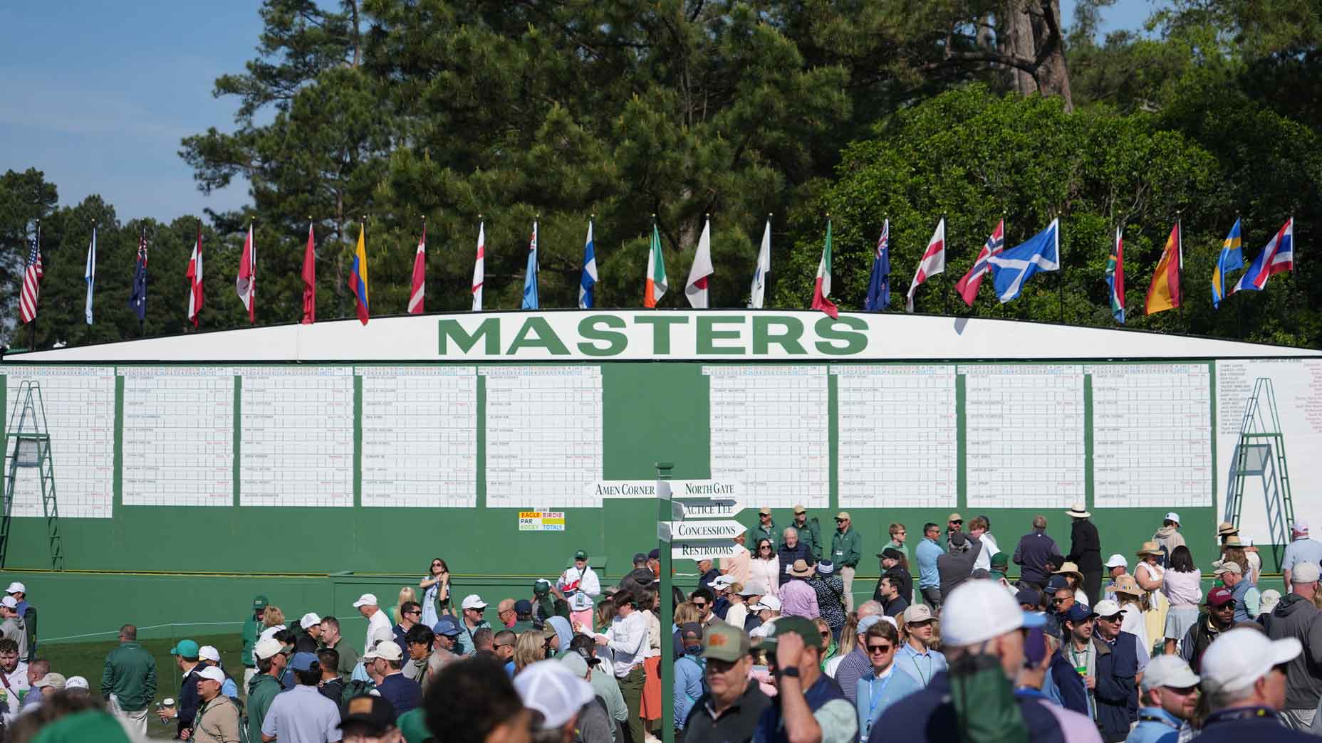A large 2025 Masters Saturday leaderboard stands outdoors with international flags on top, surrounded by a crowd of spectators in hats and golf attire. Trees frame the backdrop, capturing the excitement of this iconic tournament day.