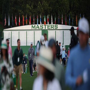 Masters patrons walk in front of a Masters leaderboard at Augusta National.
