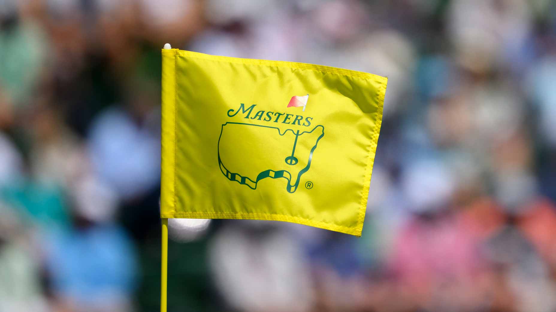Watch 2026 Masters for free: A close-up of a yellow Masters golf tournament flag featuring the United States outline and a red flag, with a blurred Masters crowd in the background.