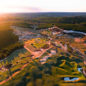 Aerial view of a golf course at sunset, with green fairways, sand traps, scattered trees, and surrounding forest under a soft orange sky.