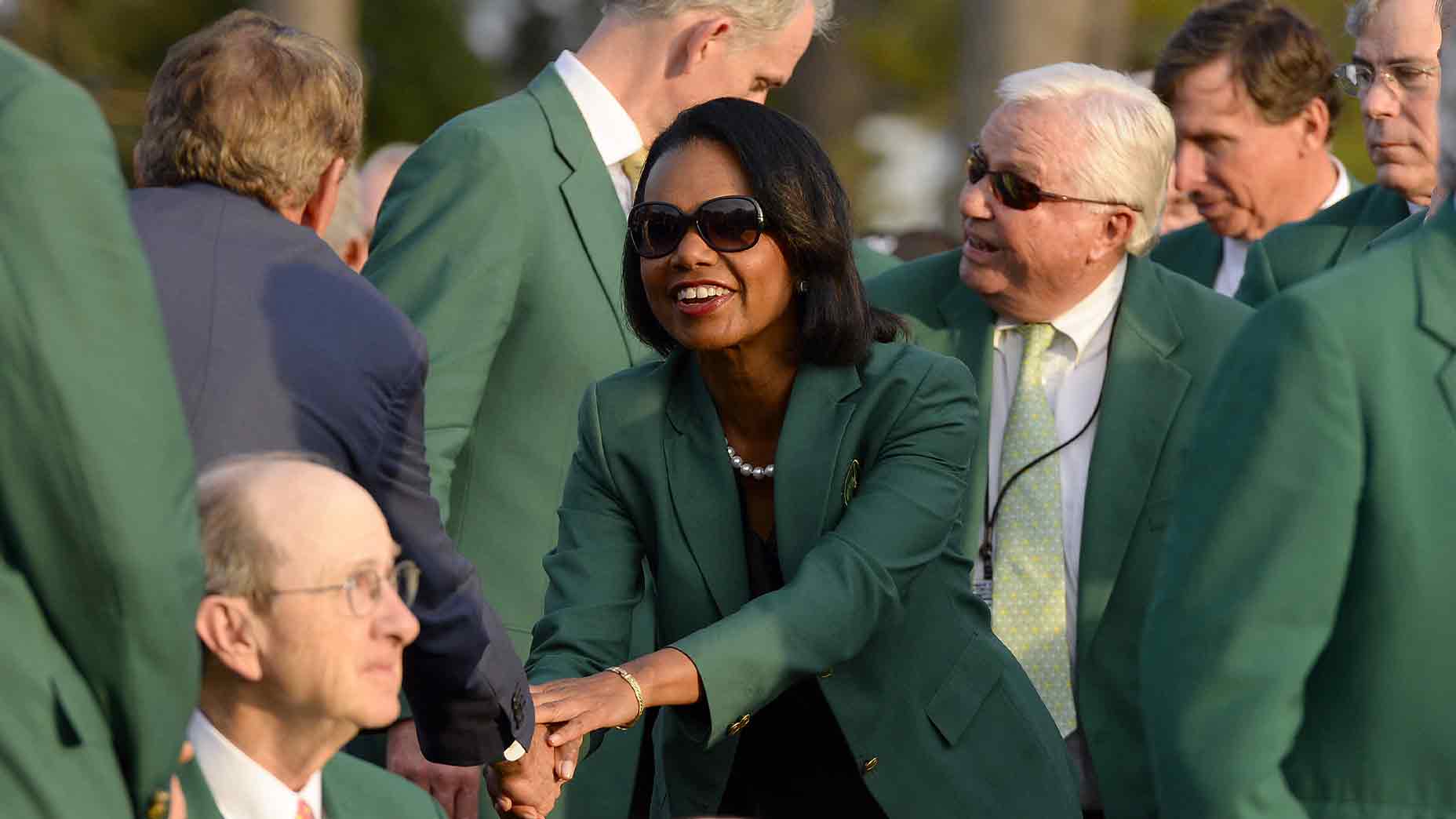 A woman wearing sunglasses and a green jacket shakes hands with a seated man, surrounded by others in green jackets at Augusta National, highlighting the history and prestige of its exclusive membership cost in an outdoor setting.
