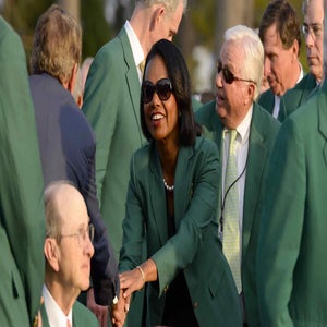 A woman wearing sunglasses and a green jacket shakes hands with a seated man, surrounded by others in green jackets at Augusta National, highlighting the history and prestige of its exclusive membership cost in an outdoor setting.