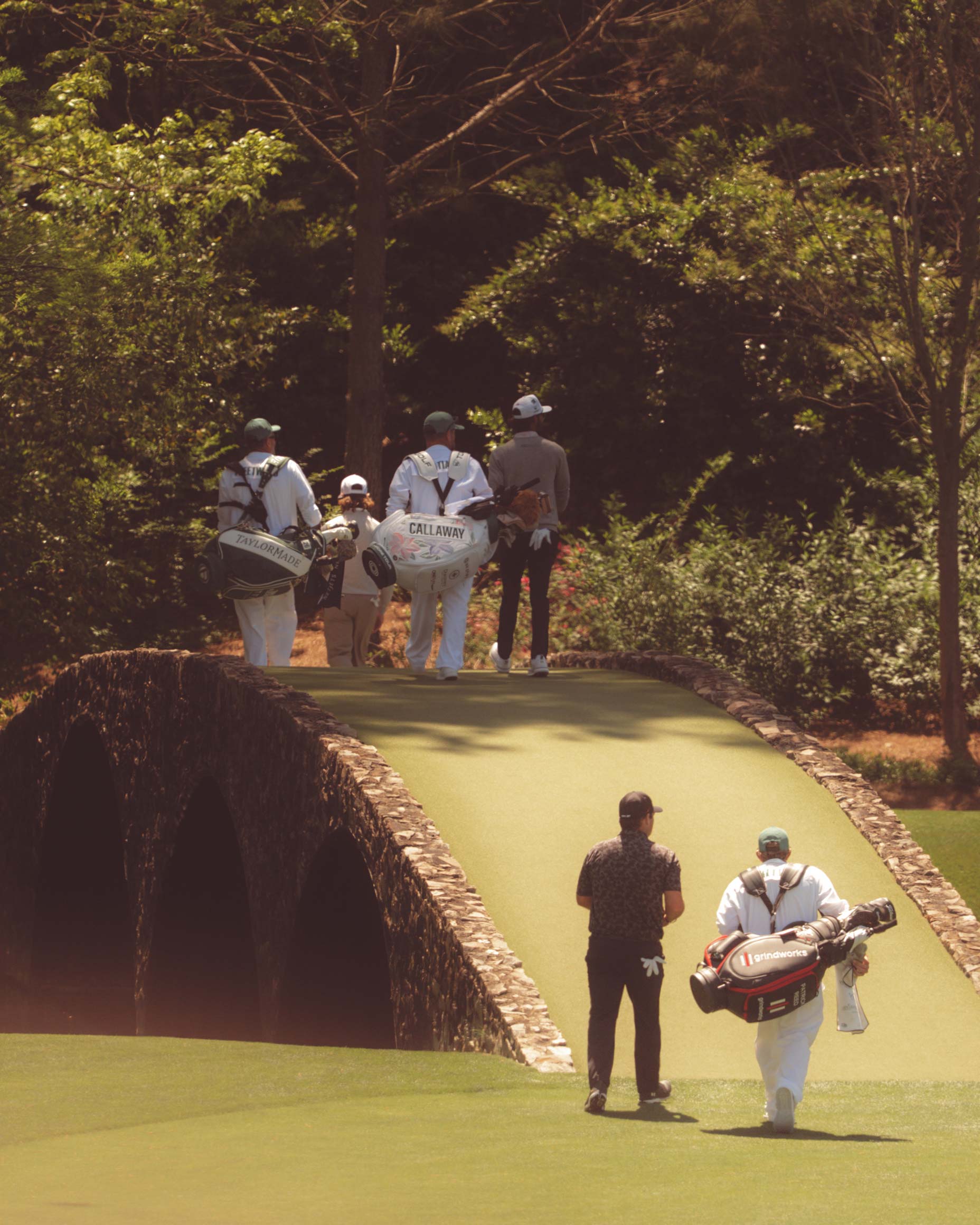 Golfers and caddies walk over a stone bridge on Augusta National’s lush golf course, surrounded by trees and greenery, with sunlight filtering through the foliage after having walked all 18.