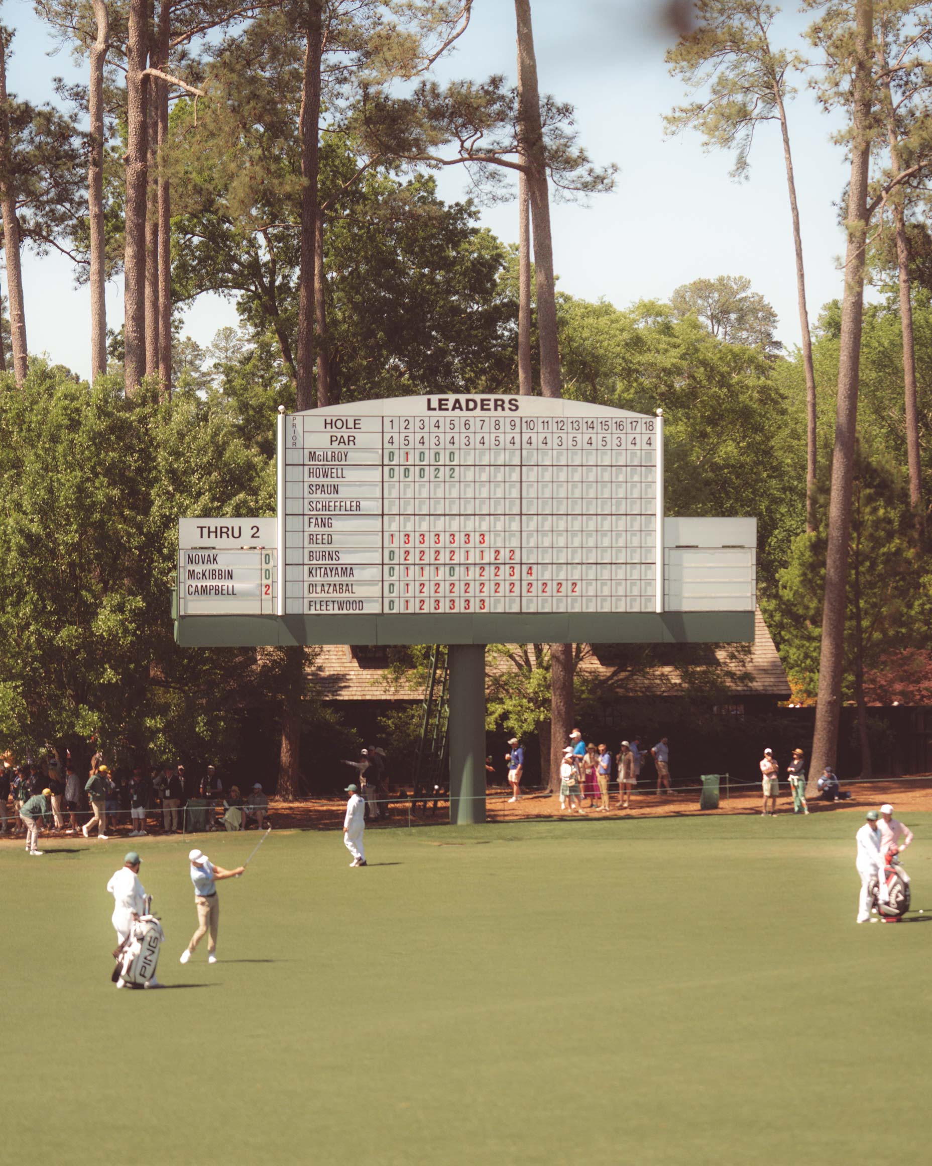 A large golf tournament leaderboard stands on a green fairway at Augusta National, surrounded by tall pine trees. Golfers and caddies who have walked all 18 are playing as spectators watch in the background.