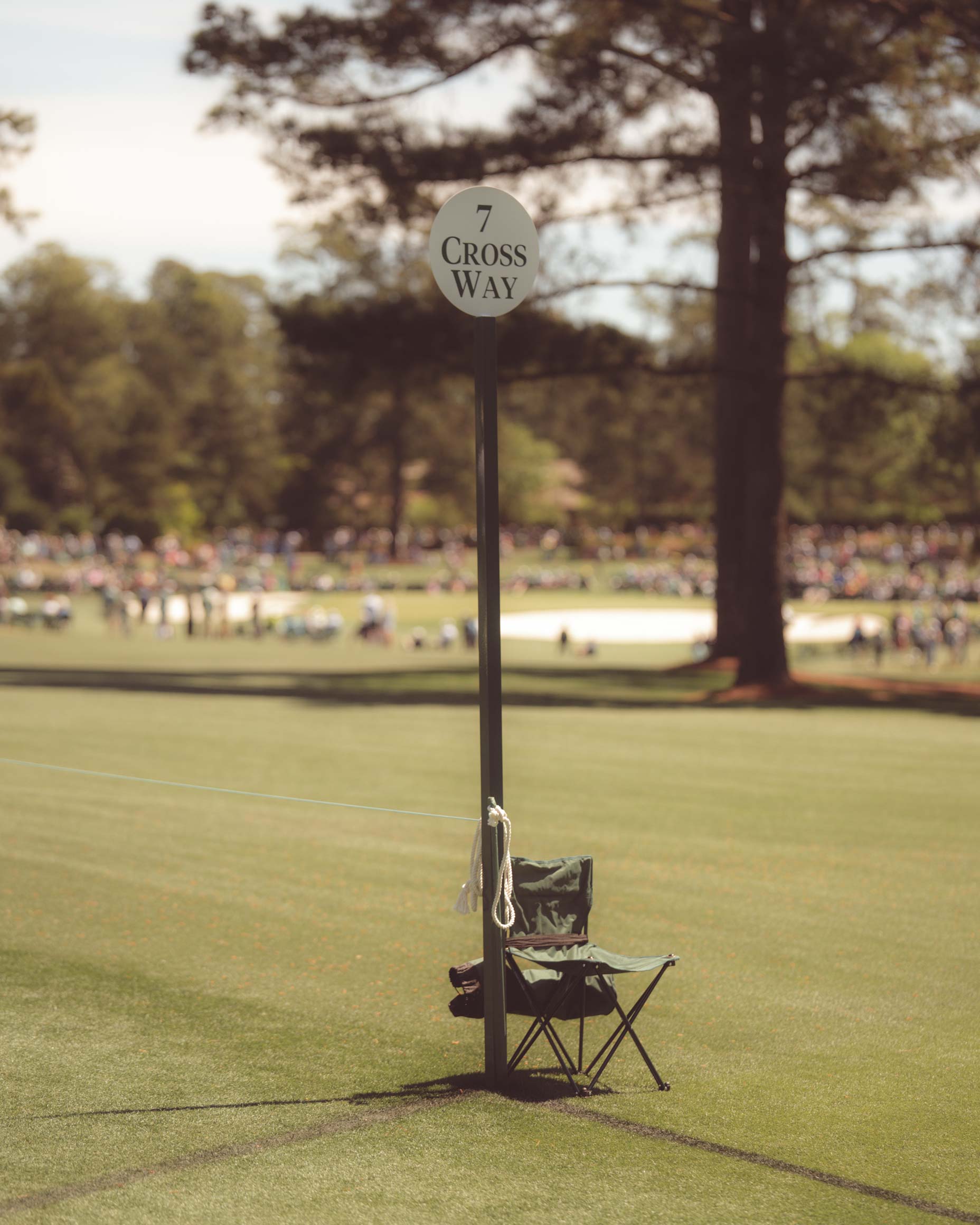 A folding chair sits next to a tall pole with a round sign reading 7 Cross Way on the grassy golf course at Augusta National, where fans who walked all 18 holes relax amid trees and a blurred crowd in the background.