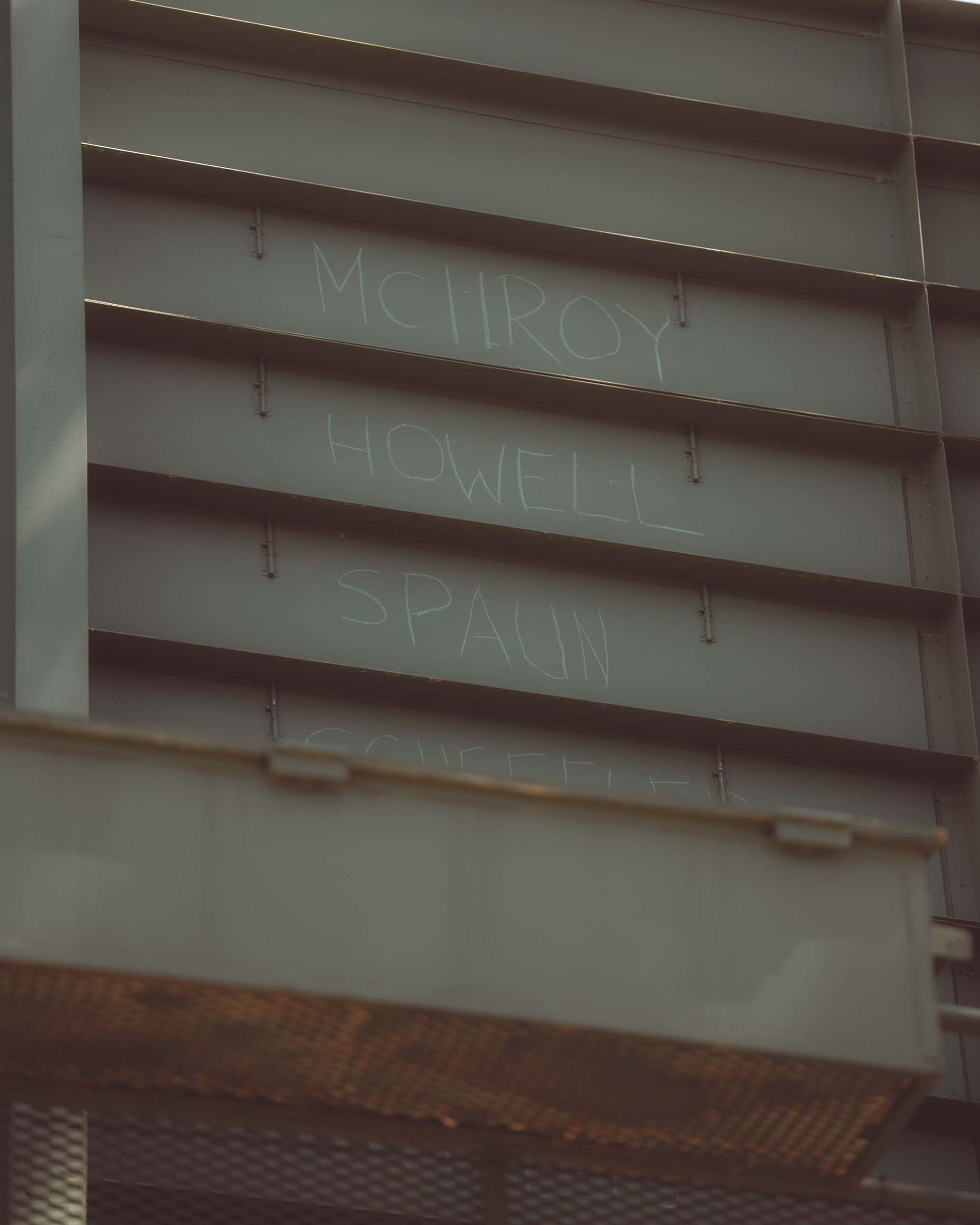 A metal wall with names MCILROY, HOWELL, and SPAUN written in white chalk across three panels—evoking the feel of Augusta National after you’ve walked all 18. A metal grate is partially visible at the bottom of the image.