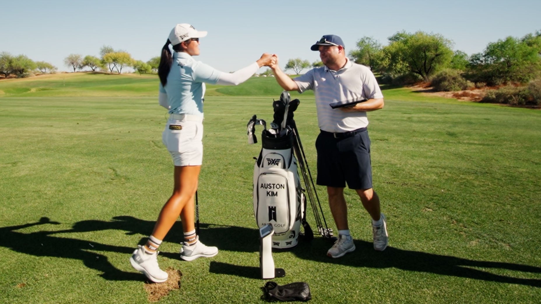 A female golfer and a male golfer fist bump next to a golf bag labeled “AUSTON KIM” on a sunny golf course. They stand on green grass surrounded by trees under a clear blue sky.