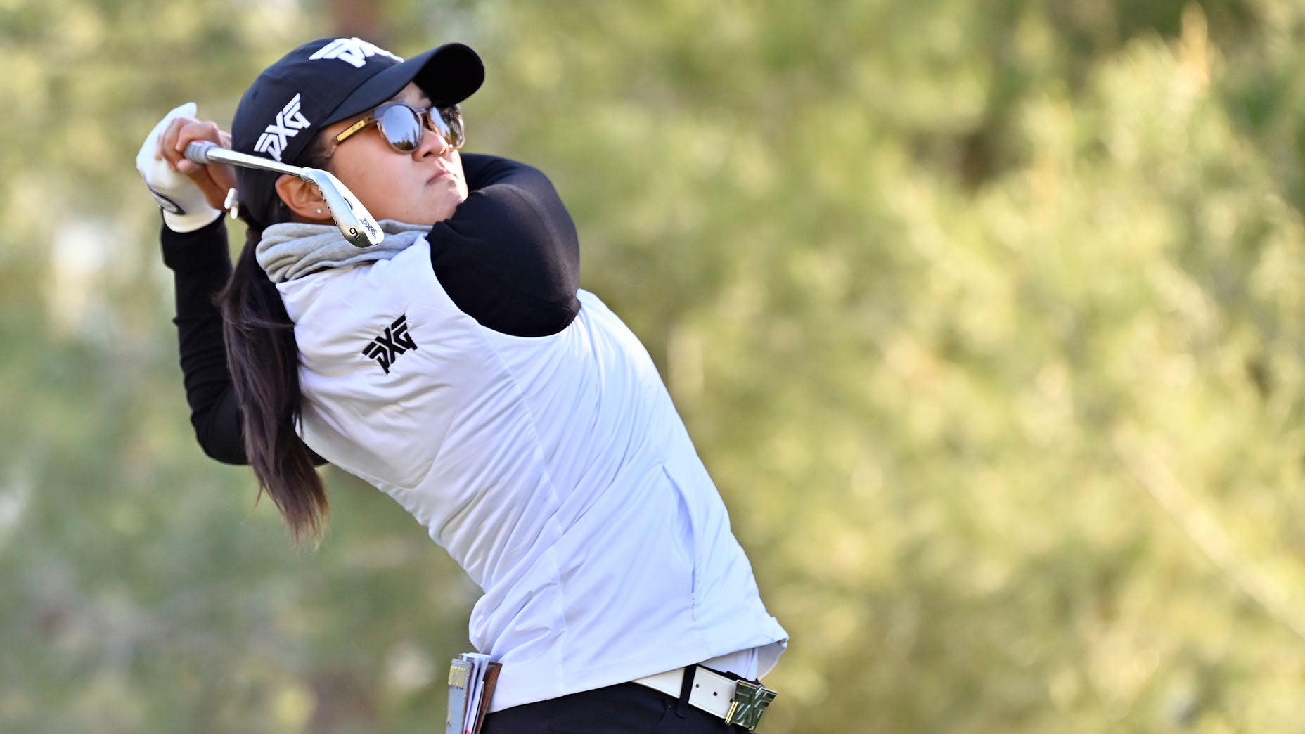 A golfer wearing sunglasses, a cap, and a white vest swings a golf club outdoors, with a blurred green background.