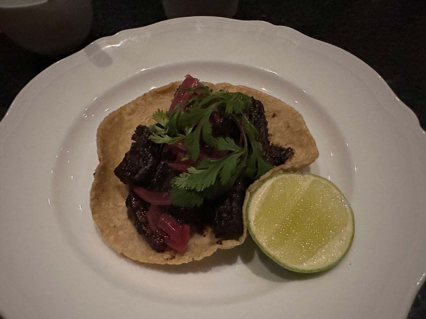 A crispy tortilla topped with dark grilled meat, pickled red onions, and fresh cilantro, served with half a lime on a white plate—perfect for enjoying delicious bites after a day at the best Mexican golf destinations.