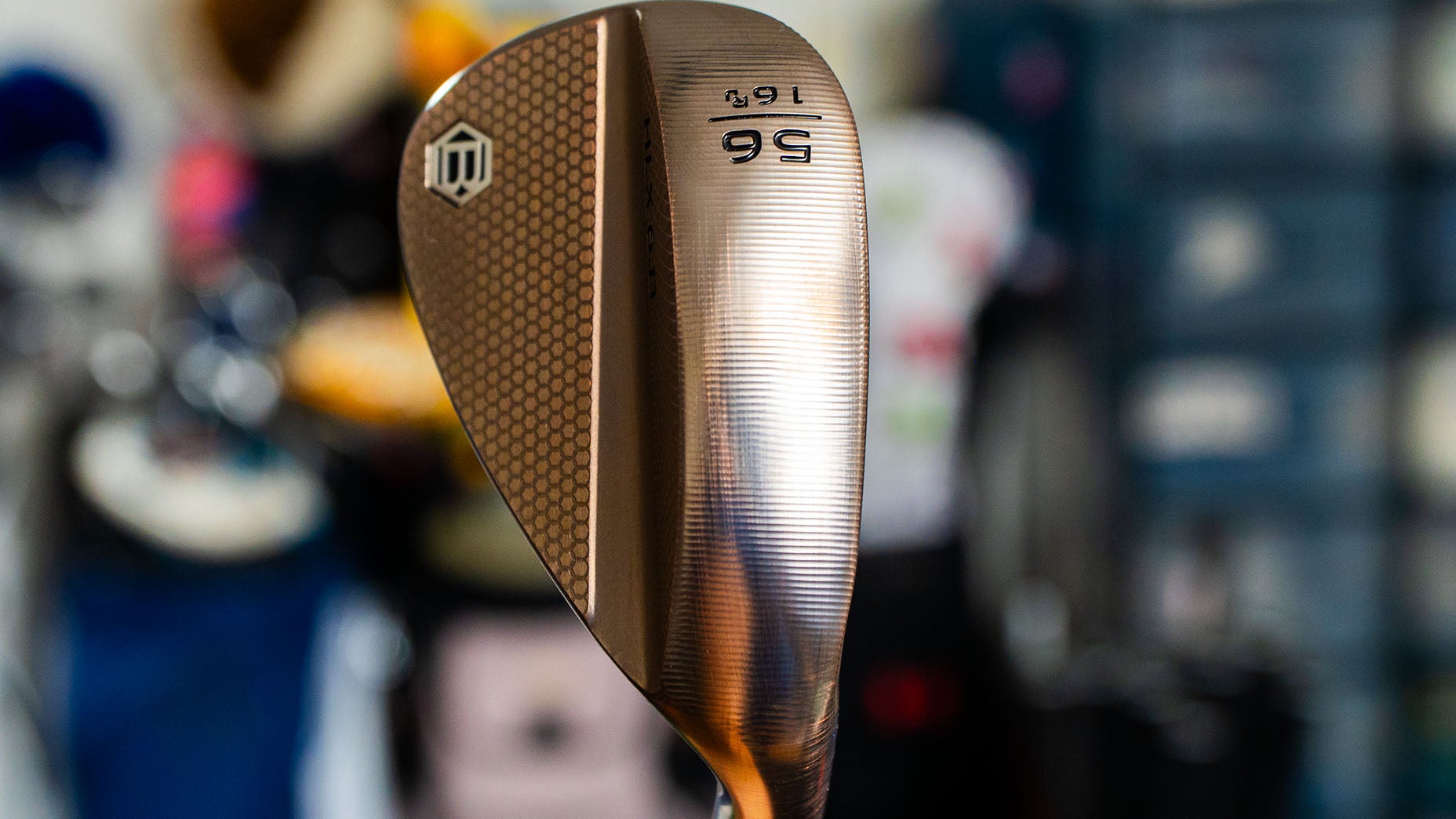 Close-up of a golf wedge with a bronze finish, showing the numbers 56 and 16 engraved on the club head, along with a logo on the textured surface. The background is blurred.