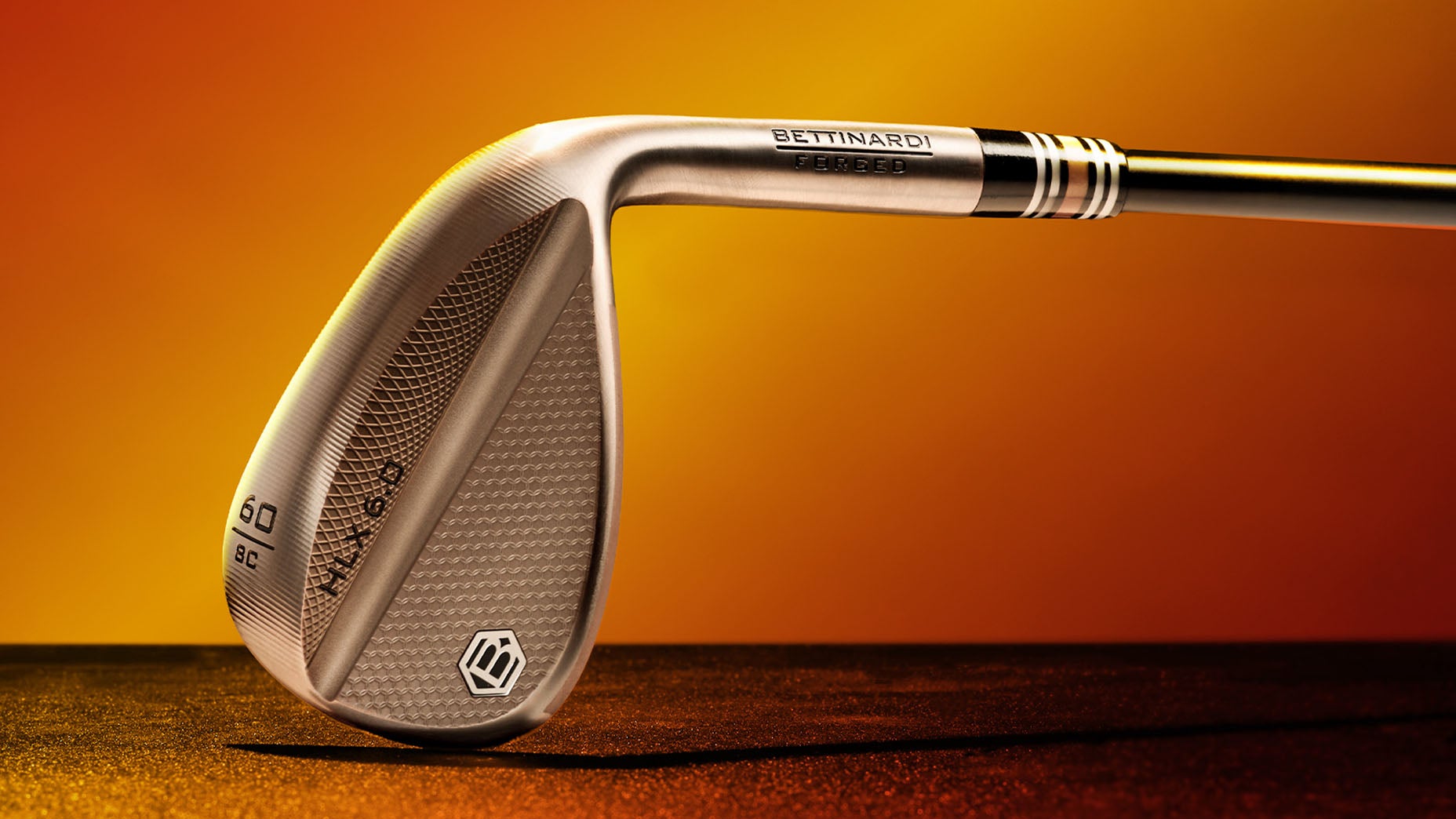 Close-up of a Bettinardi forged golf wedge with a metallic finish, engraved logo, and textured face, set against a gradient orange and yellow background.