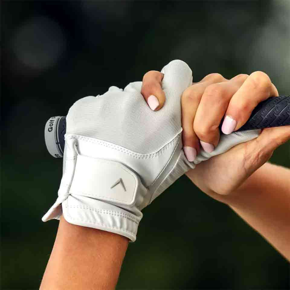 A close-up of two hands gripping a golf club — one with a white glove, the other with pale pink manicured nails — captures a special Mother's Day moment. The background is softly blurred, emphasizing this intimate gesture.