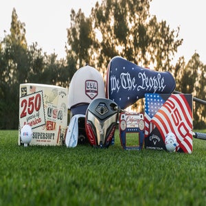A collection of patriotic-themed golf accessories, including golf balls, a cap, golf club head covers, and boxed sets, displayed on grass with trees in the background at sunset.