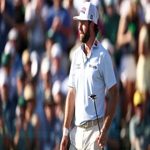 Cameron Young, wearing a white cap and striped shirt, holds a putter on the golf course during the Masters, with a blurred crowd in the background.