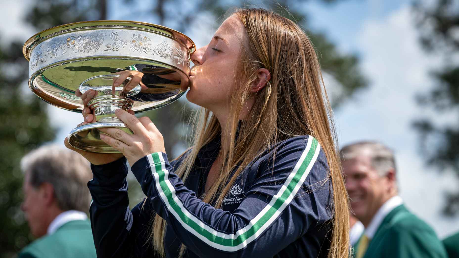 A young woman with long hair kisses a large silver trophy, celebrating her Augusta National Women's Amateur victory.