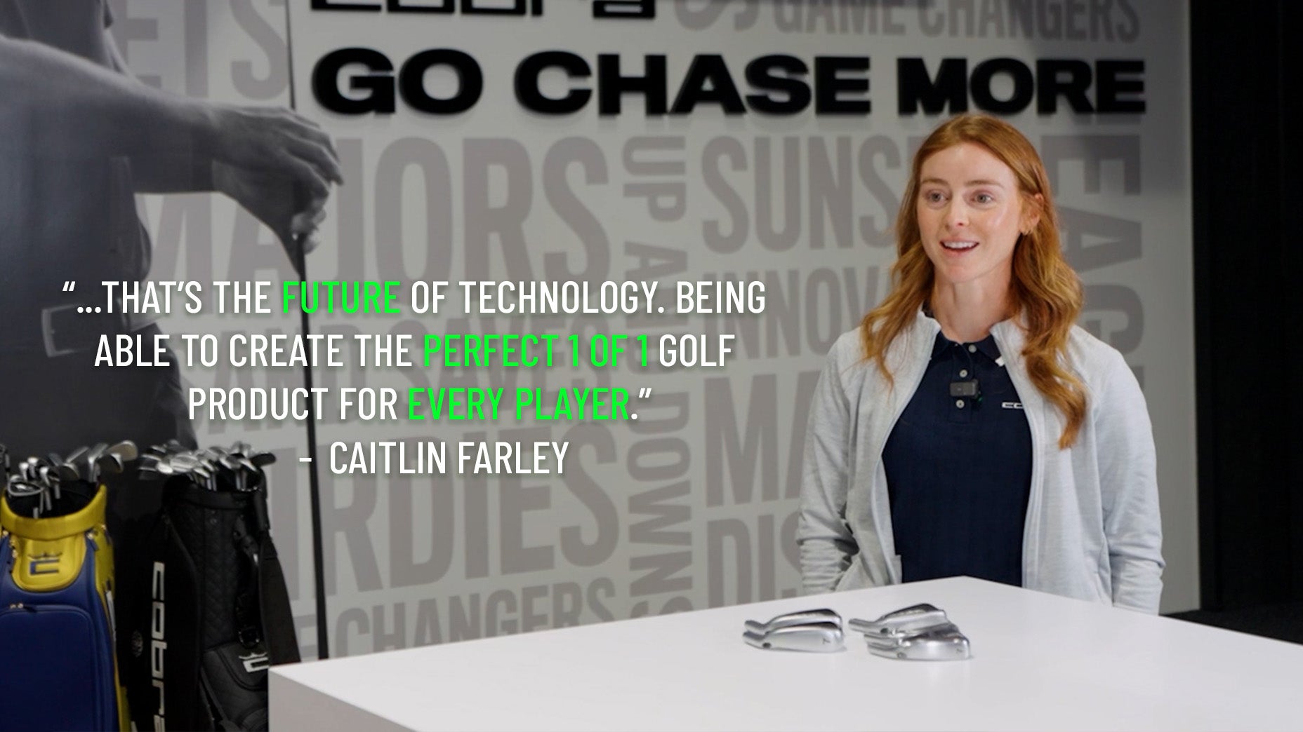 A woman with long red hair sits at a white table with two Cobra 3DP Irons golf club heads on it, smiling. Beside her is a rack of golf clubs. A quote about golf technology is displayed on the wall behind her.