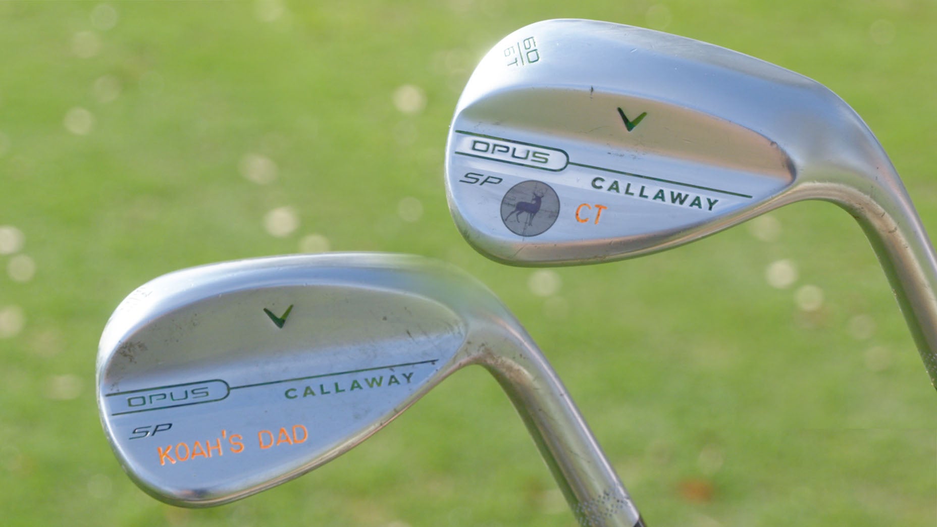 Two silver Callaway golf clubs with custom engravings on their heads rest upright on green grass. One club is labeled KOAHS DAD in orange lettering. Both clubs show the Opus and Callaway branding.