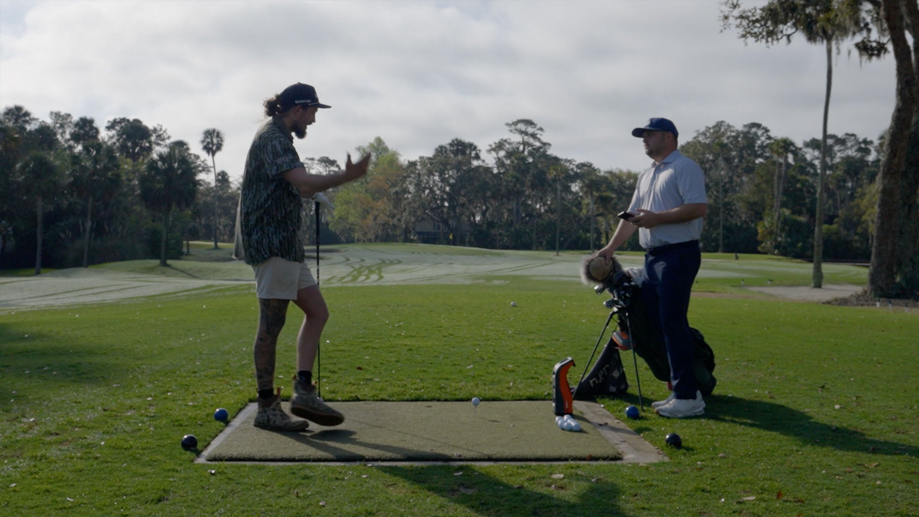 Two men stand on a golf course tee box; one, in shorts and a patterned shirt, gestures animatedly, while the other, in golf attire, stands near a golf bag in a relaxed stance. Trees and fairways are visible in the background.