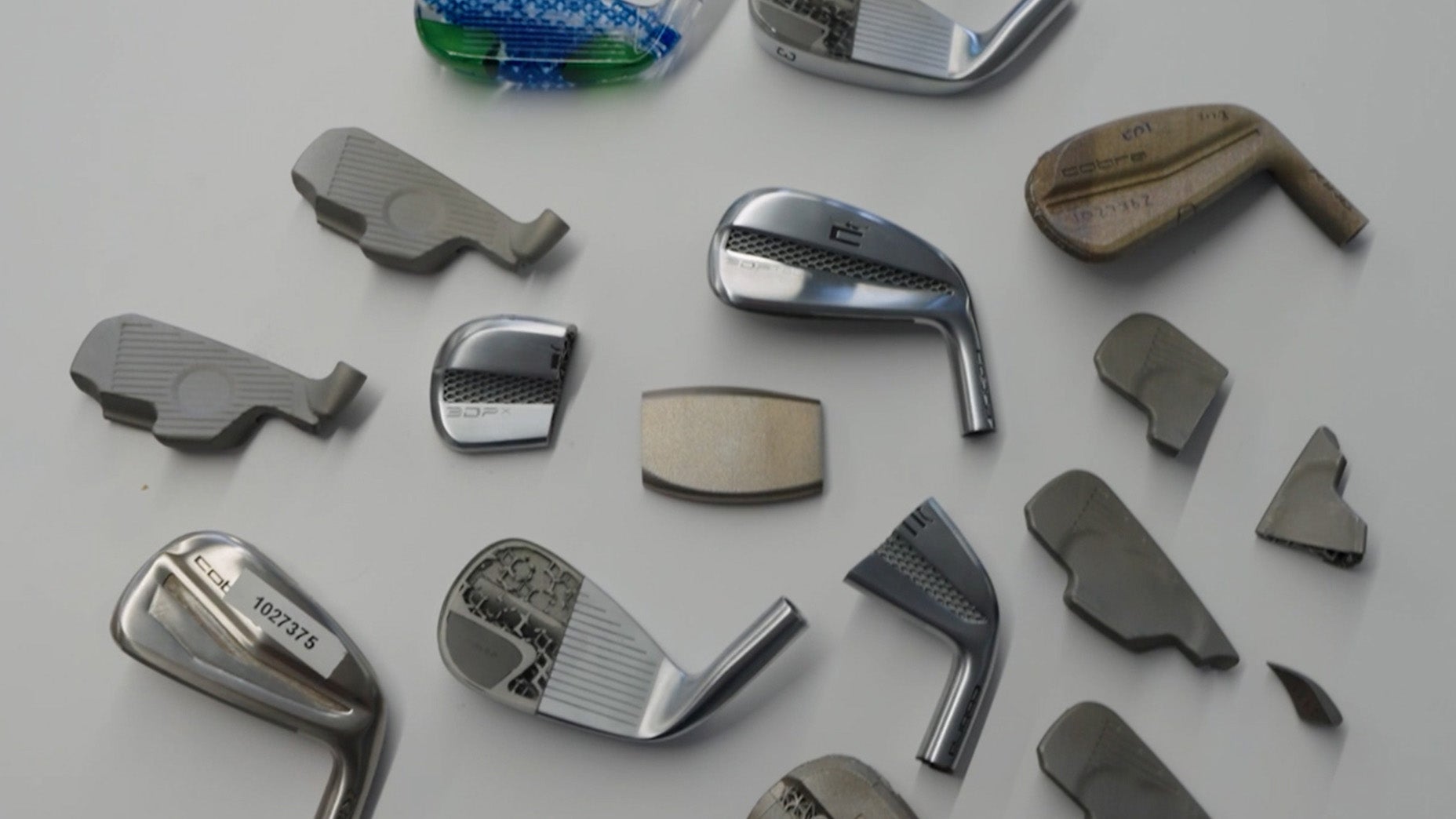 Various stages of Cobra 3DP Irons club heads are arranged on a light surface, showcasing unfinished, partially finished, and polished metal pieces in different shapes and sizes.