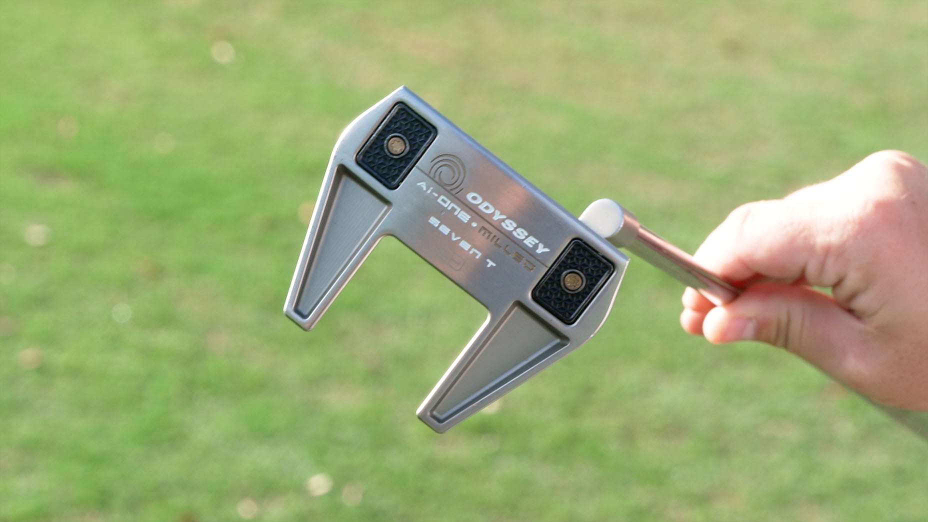A close-up of a hand holding an Odyssey Arm Lock Seven T golf putter, with a grassy area in the blurred background.