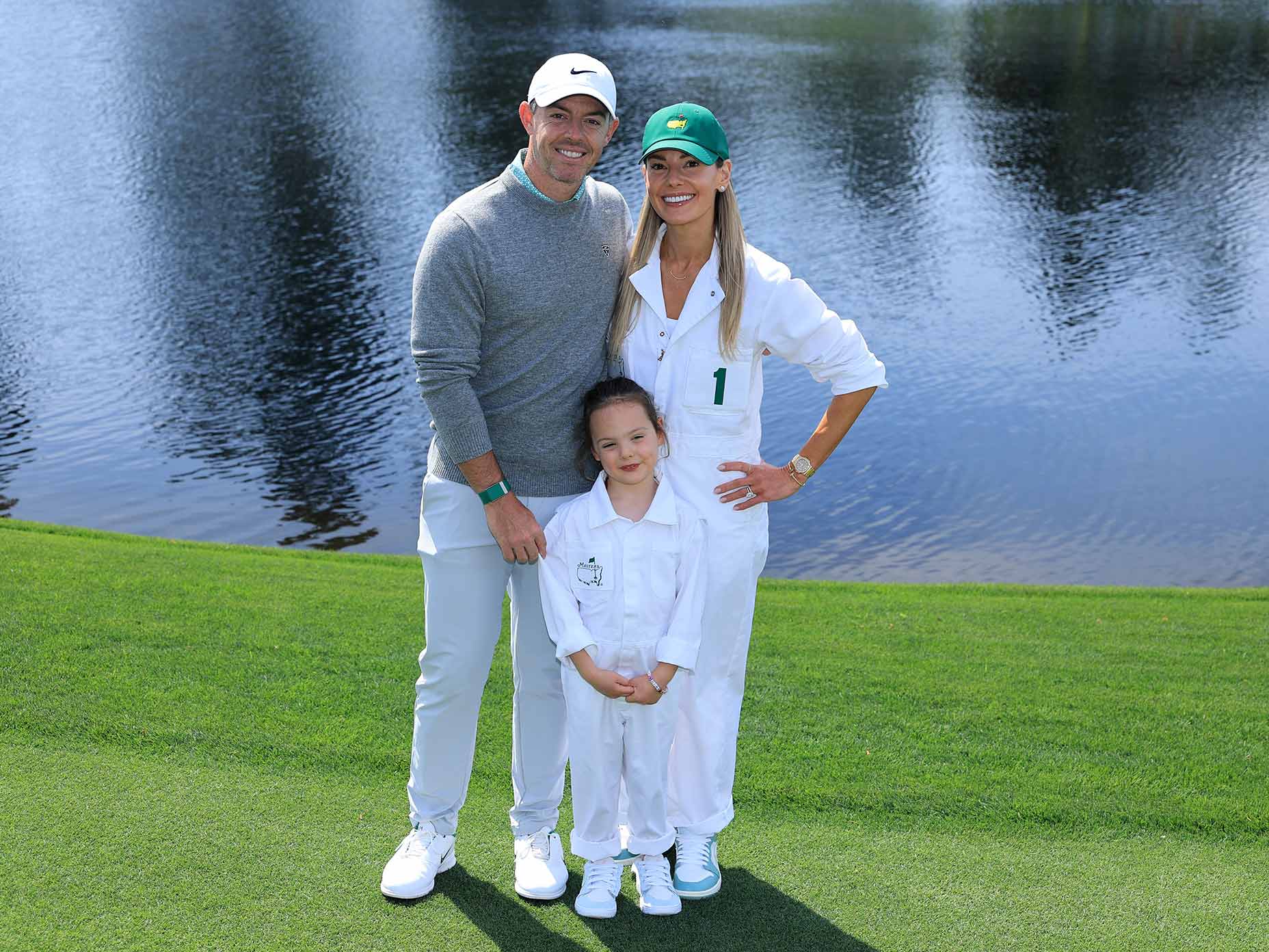 A man in a gray sweater and white pants stands on the grass beside a woman and a child dressed in white jumpsuits and green hats, all smiling in front of a pond at the Masters par 3 course.