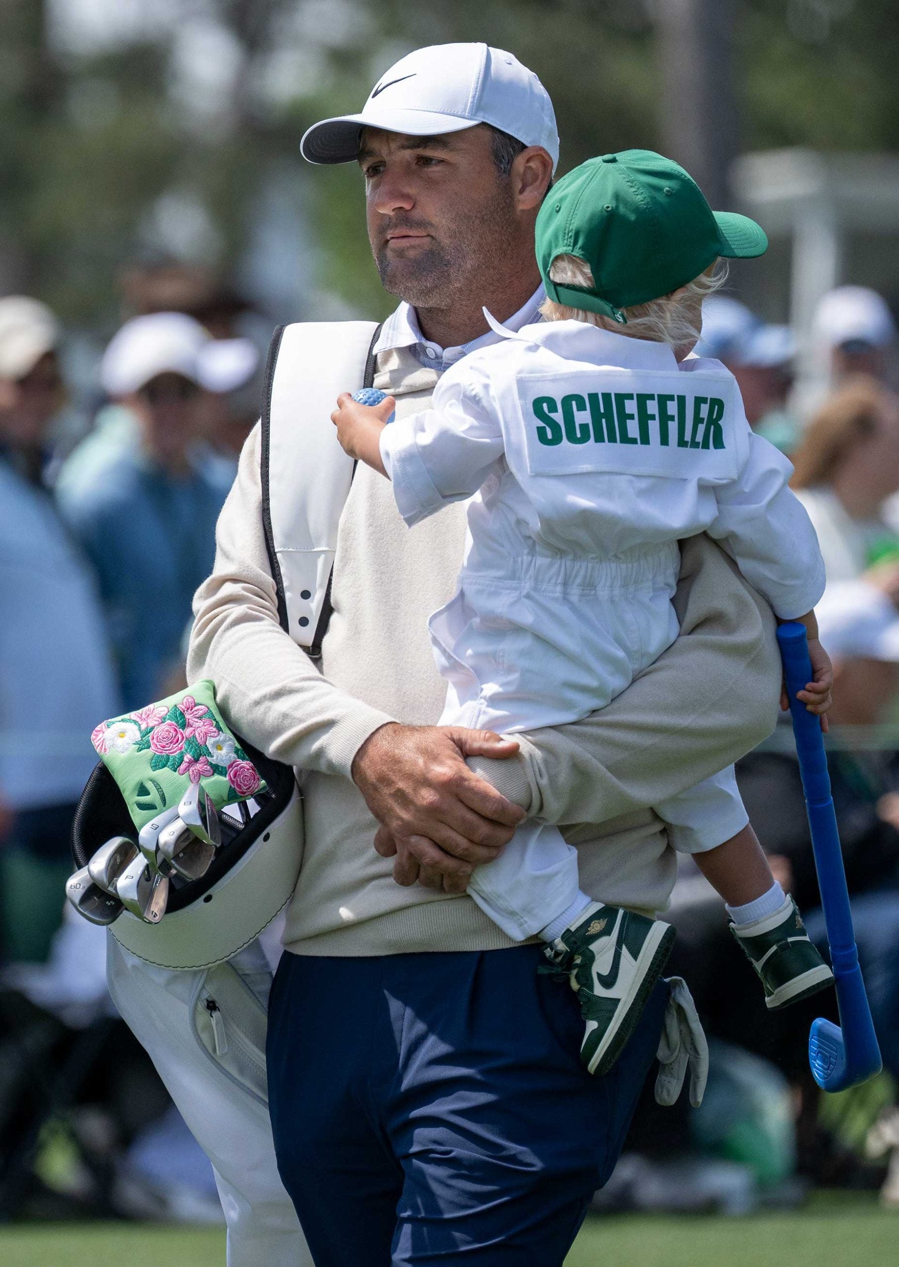 A golfer in a white cap and beige sweater carries a young child dressed in white with 