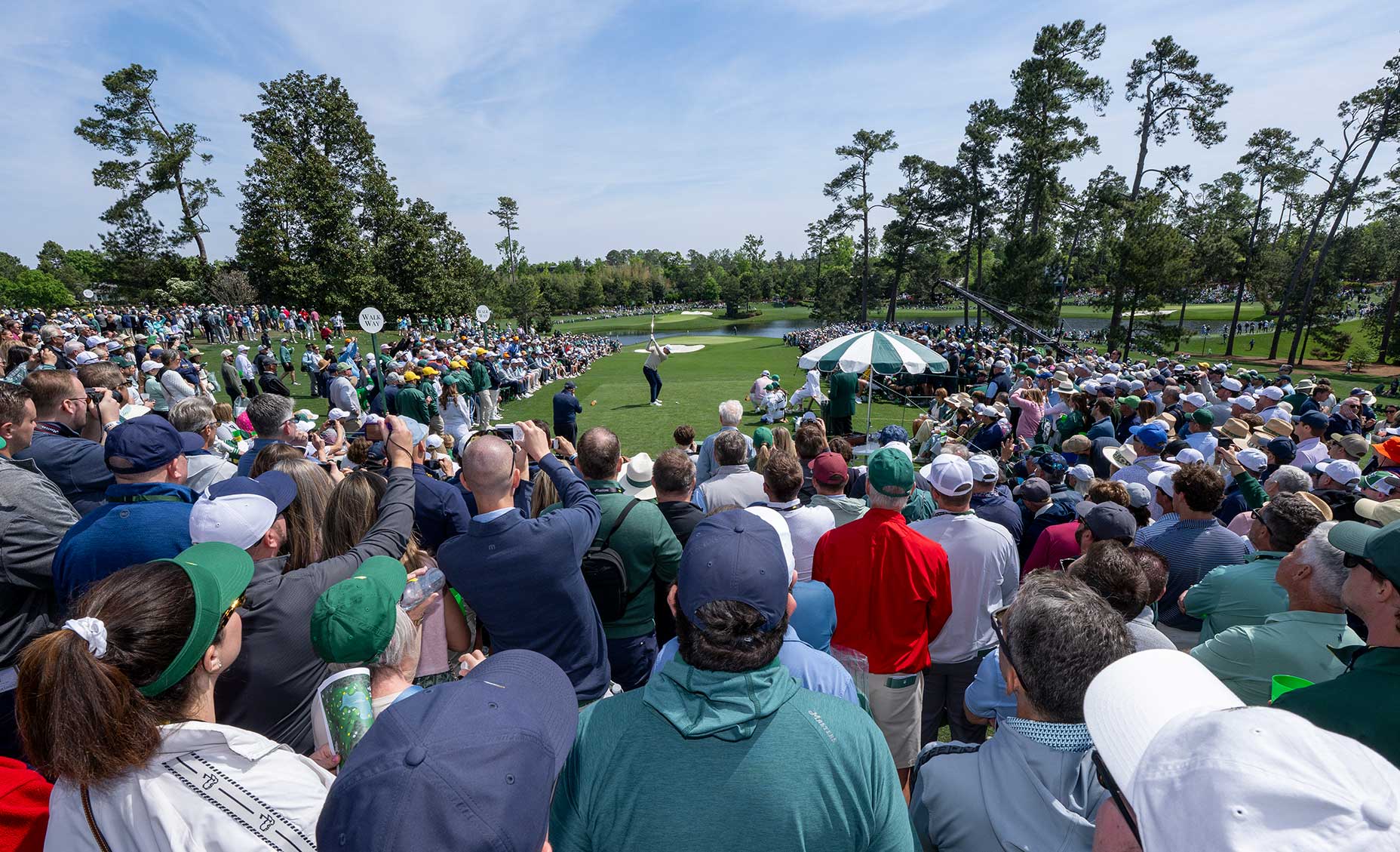 A large crowd gathers around a golf tee at the Masters, watching as a golfer prepares to swing on the lush par 3 course. Tall trees frame the scene under a clear, sunny sky, with spectators dressed in casual clothing and hats.
