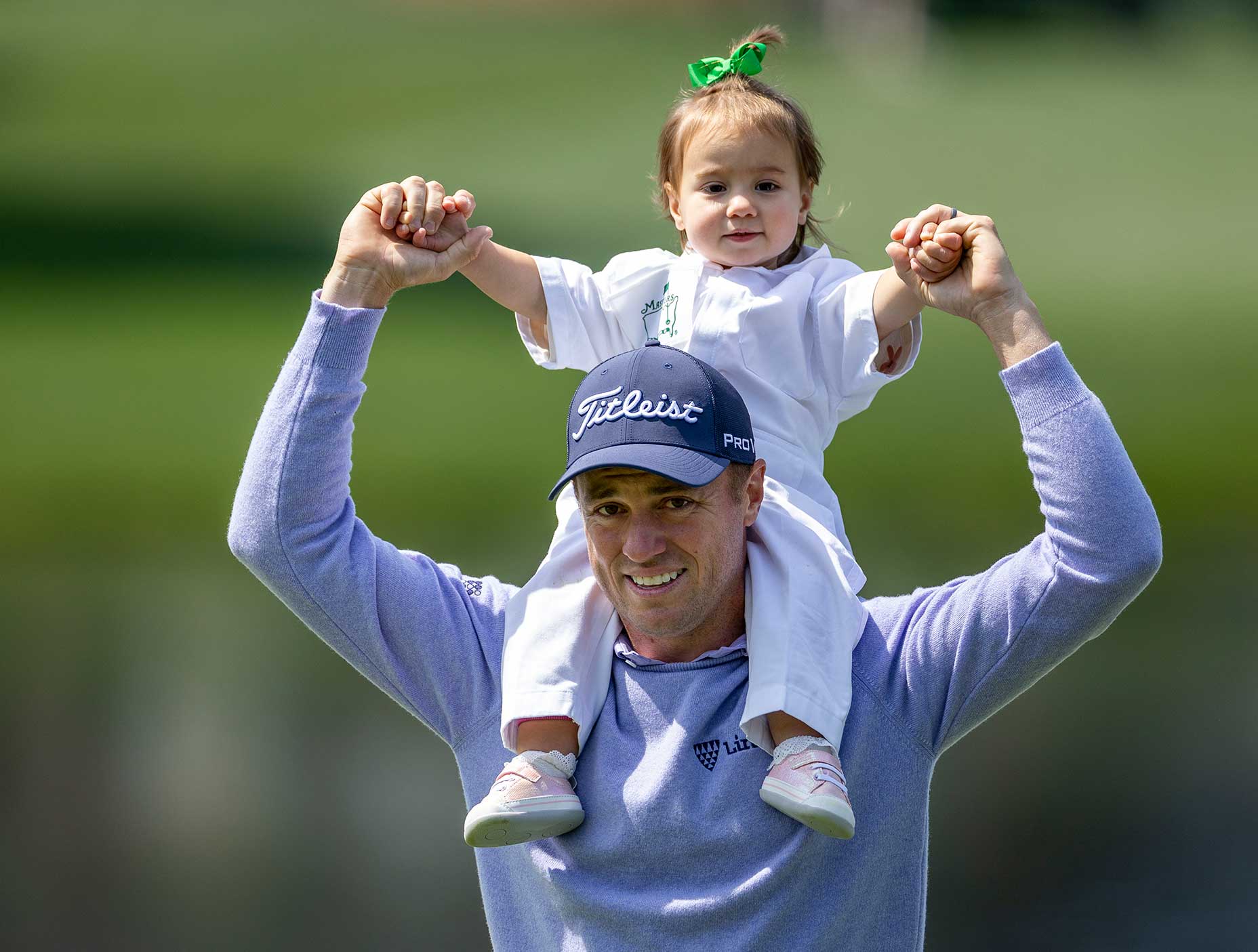 A smiling man wearing a cap holds a young child with a green bow on his shoulders, both holding hands and enjoying time outdoors on a golf course during the Masters par 3 event.