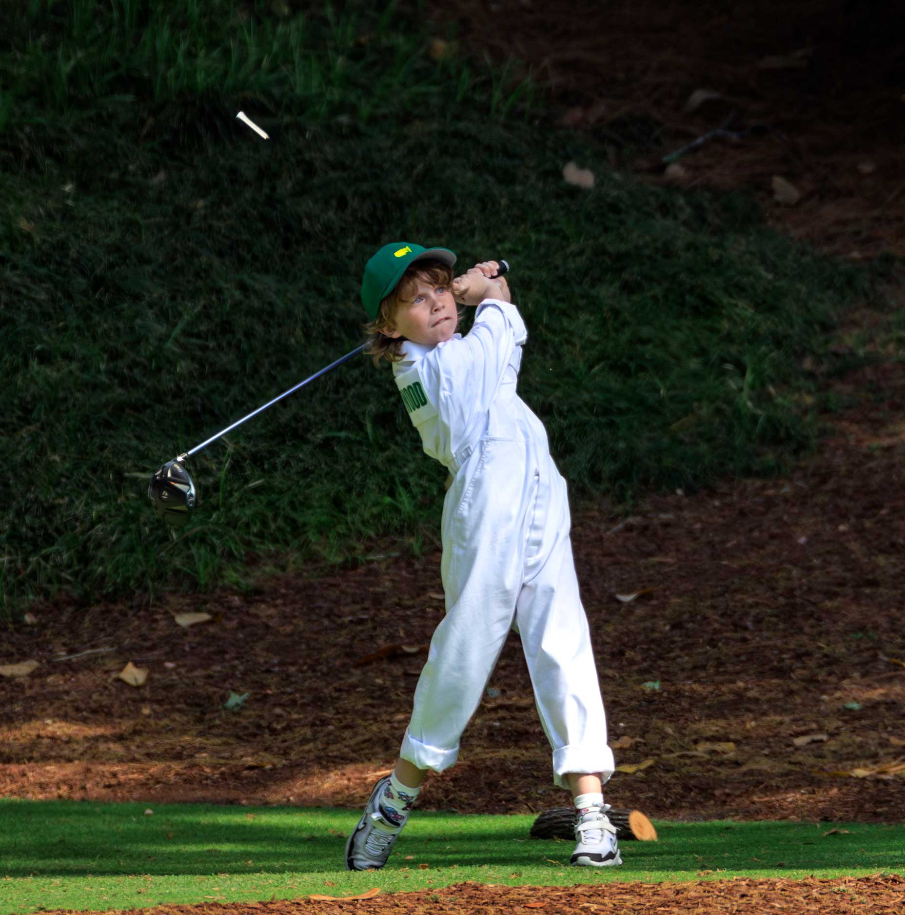 A young child in a green cap and white jumpsuit swings a golf club on a par 3, with dirt flying in the air and greenery reminiscent of the Masters in the background.