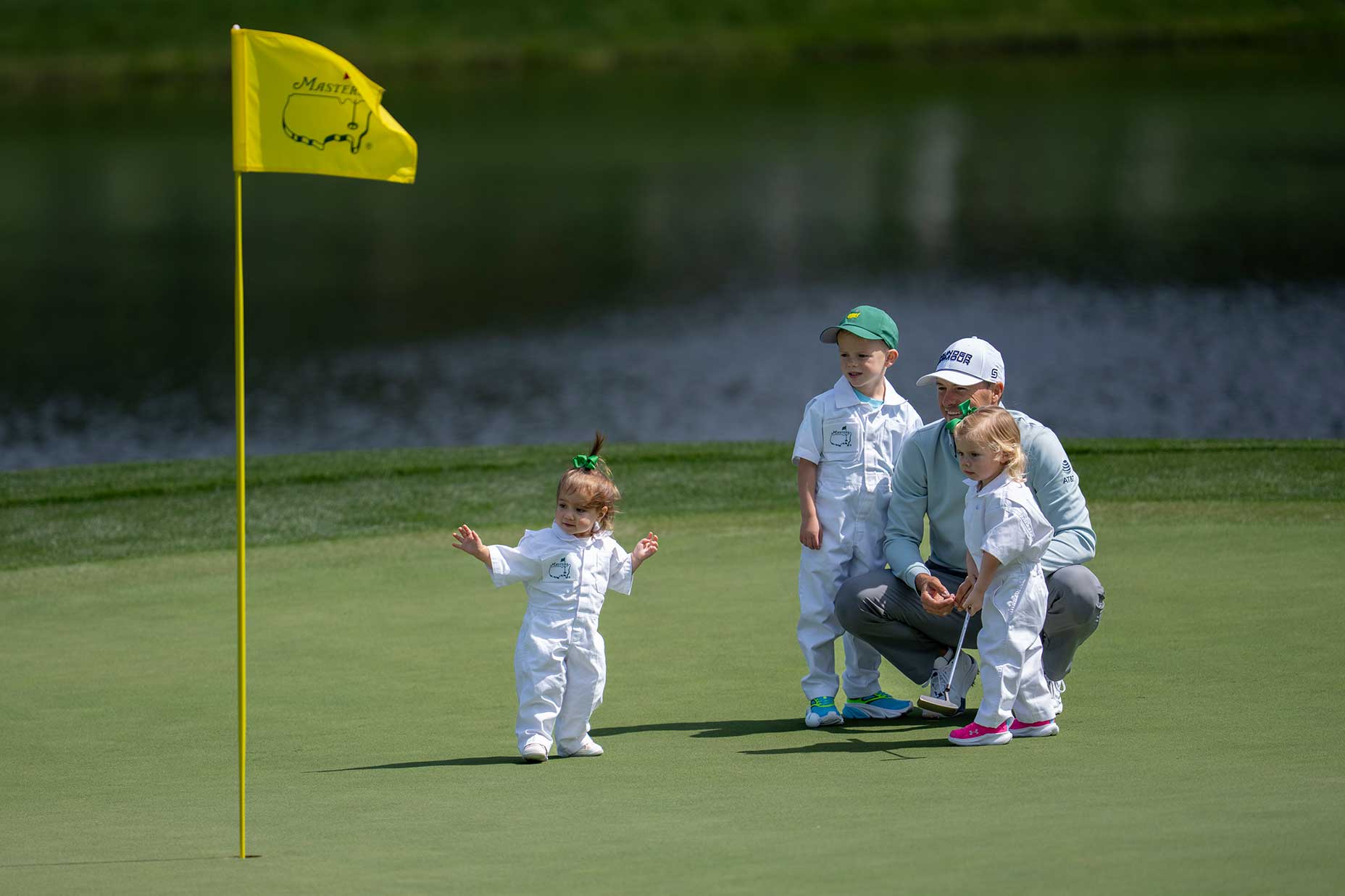 A man and three young children in white jumpsuits stand on a Masters par 3 golf green near a yellow flag, with water and grass in the background. One child walks ahead as the others watch.